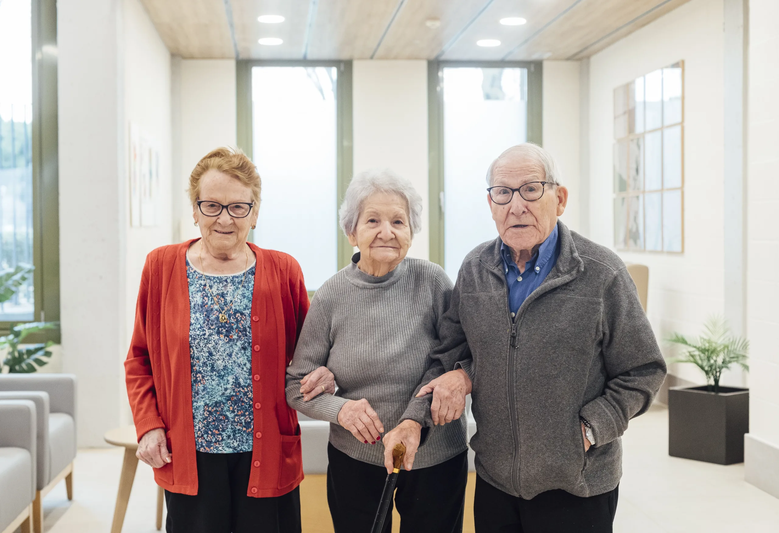 Dos señoras y un señor mayor cogidos de la mano mientras pasean por los pasillos de la residencia